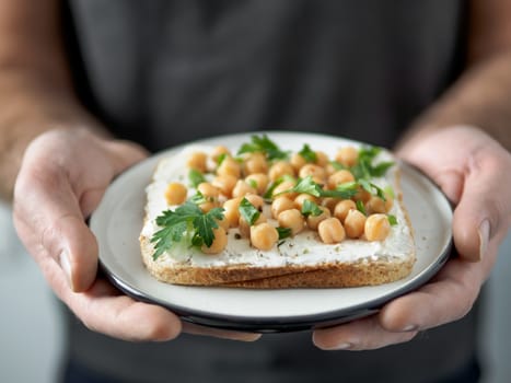 Hands takes plate with vegan sandwich. Healthy appetiezer - whole wheat bread toast with plant-based soft cheese, chickpea and fresh green parsley. Vertical