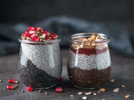 Two chia pudding in glass jars on dark table. Two colors charcoal chia puding with pomegranate, almond, hemp seeds. Chocolate chia pudding with jam and granola. Copy space. Healthy overnight breakfast