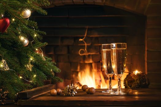 Christmas composition - Two glasses with champagne on a wooden table near a Christmas tree in a room with a burning fireplace.