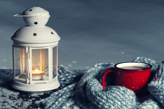 Lantern with a burning candle and a red mug with hot coffee on a snowy wooden table.