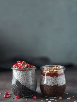 Two chia pudding in glass jars on dark table. Two colors charcoal chia puding with pomegranate, almond, hemp seeds. Chocolate chia pudding with jam and granola. Copy space. Healthy overnight breakfast