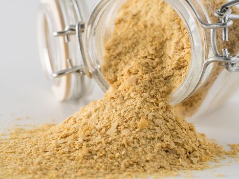 Nutritional yeast background. Nutritional inactive yeast spilled from glass jar on white background. Copy space.Side view.Nutritional yeast is vegetarian superfood with cheese flavor, for healthy diet