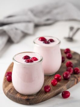 Yogurt with cranberry. Two glass jar with pink yoghurt or milkshake and red cranberries on wooden cutting board over white marble background. Copy space for text. Vertical.