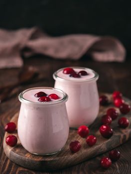 Yogurt with cranberry. Two glass jar with pink yoghurt or milkshake and red cranberries on wooden cutting board over dark background. Copy space for text. Vertical.