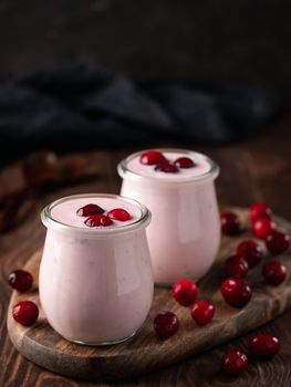 Yogurt with cranberry. Two glass jar with pink yoghurt or milkshake and red cranberries on wooden cutting board over dark background. Copy space for text. Vertical.