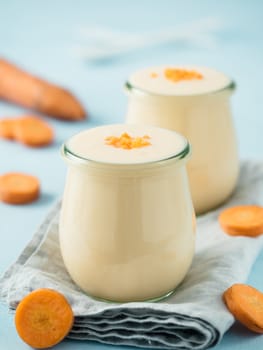 Yogurt with carrot. Vegetable yogurt. Two glass jar with yellow orange yoghurt or milkshake and fresh carrot on blue background. Copy space for text. Vertical.