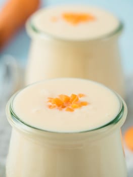 Yogurt with carrot. Vegetable yogurt. Two glass jar with yellow orange yoghurt or milkshake and fresh carrot on blue background. Copy space for text. Vertical.