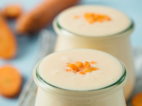 Yogurt with carrot. Vegetable yogurt. Two glass jar with yellow orange yoghurt or milkshake and fresh carrot on blue background. Copy space for text