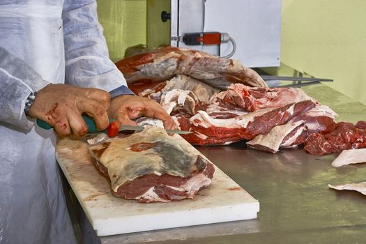 butcher in his butcher's shop to eliminate the excess fat from a large meat cut by a special knife. Hands and stools dirty with blood.