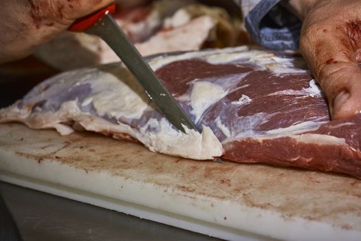 butcher who using a knife is slicing the meat in order to be sold and later eaten in his butcher's shop.