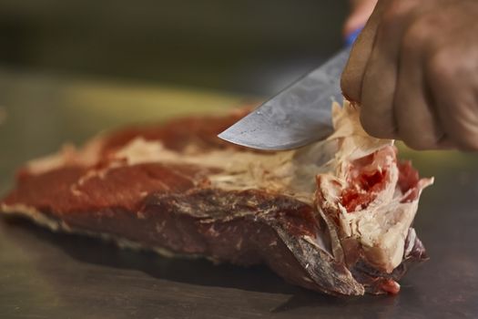 butcher who using a knife is slicing the meat in order to be sold and later eaten in his butcher's shop.