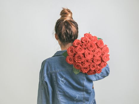 Back view of young woman in denim jacket holding bunch of Living Coral roses on shoulder. Girl with bun updo in jeans holding flowers in Color of Year 2019 Living Coral. Copy space.