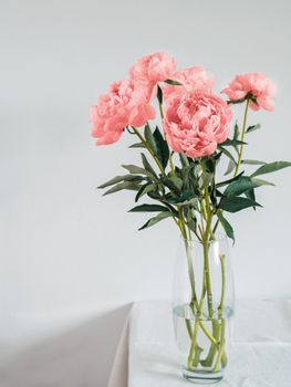 Beautiful pink Living Coral peony bouquet in glass vase on table with white linen tablecloth. Vertical. Copy space for text. Toned filter