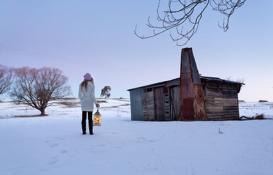 A woman standing in a snow covered field with timber shed or stable.  She is wearing a sweater dress with long boots and is holding a lantern in the coolness of dusk