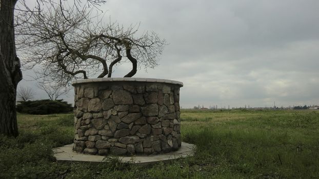 Panorama with a well made of stones in a rural landscape with cloudy sky: symbol of solitude and melancholy.