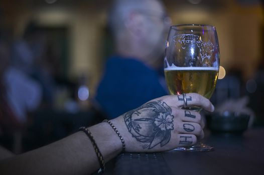 Tattooed hand holding a glass of beer at a table in a bar in the night.