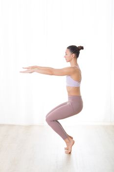 Portrait of gorgeous active sporty young woman practicing yoga in studio. Beautiful girl practice Utkatasana, awkward yoga pose. Healthy active lifestyle, working out in gym.