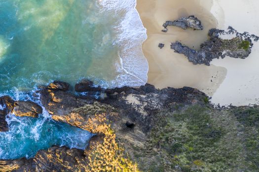 Waves wash onto the beach and  into an eroded rock tunnel in the cliffs of the headland. High above a secret window to peek down inside