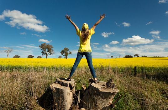 Woman enjoys the countryside views standing on large tree stump to view the canola fields in flower during spring.