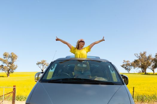 Happy smiling woman standing out the sky roof of her 4wd car by the farm gate of flowering canola fields in rural Australia