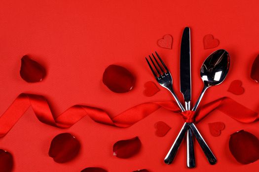 Cutlery set tied with silk ribbon rose flower petals and hearts on red background Valentine day dinner concept