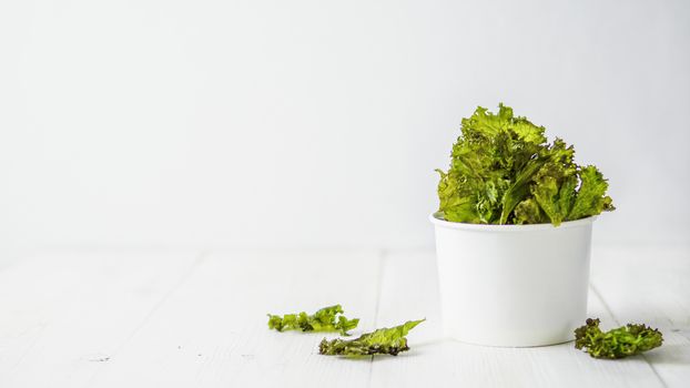 Kale Chips with salt in paper cup. Homemade healthy snack for low carb, keto, low calorie diet. White wooden background. Ready-to-eat kale chips, copy space for text. Banner