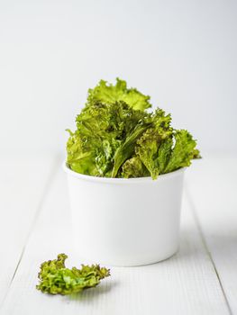 Kale Chips with salt in paper cup. Homemade healthy snack for low carb, keto, low calorie diet. White wooden background. Ready-to-eat kale chips, copy space for text. Vertical