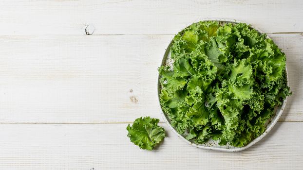 Kale close up. Green vegetable leaves, top view on white craft plate over white wooden tabletop. Healthy eating, vegetarian food,dieting concept. Top view or flat lay. Copy space. Health kale benefits