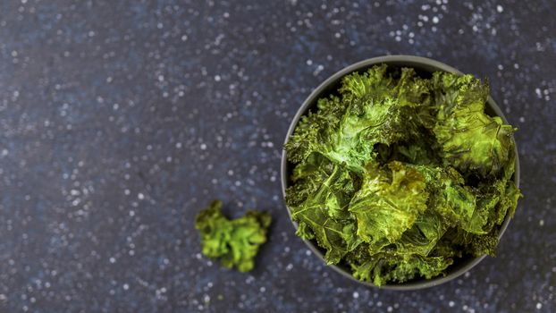 Green Kale Chips with salt in bowl. Homemade healthy snack for low carb, keto, low calorie diet. Dark blue background. Ready-to-eat kale chips, copy space for text. Top view or flat lay. Banner