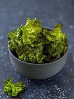 Green Kale Chips with salt in bowl. Homemade healthy snack for low carb, keto, low calorie diet. Dark blue background. Ready-to-eat kale chips, vertical