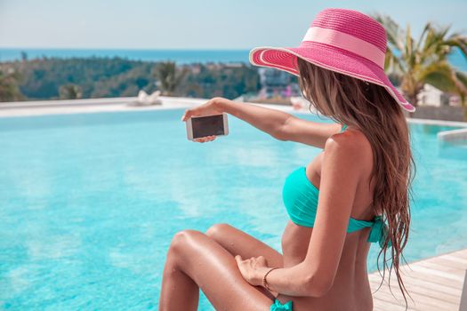 Portrait of a beautiful woman in bikini and sunhat making selfie photo on smartphone outdoors sitting at tourist resort by the pool