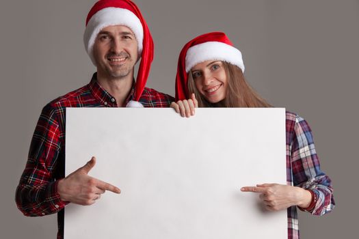 Happy couple in Santa hats holding blank banner and pointing at copy space