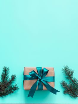 Gift box in craft wrapping paper and green satin ribbon on turquoise blue background, copy space top. Beautiful Christmas or New Year present and fir branches, flat lay or top view. Vertical