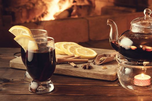 Two glasses of mulled wine and teapot on a wooden table in front of a burning fireplace.