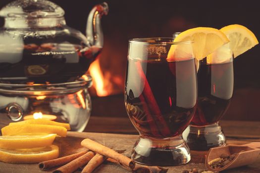 Two glasses of mulled wine and teapot on a wooden table in front of a burning fireplace.