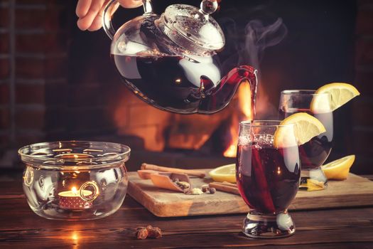 Pouring mulled wine into glasses from a teapot in a room with a fireplace.