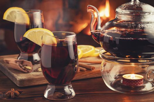 Two glasses of mulled wine and teapot on a wooden table in front of a burning fireplace.