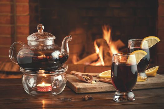 Two glasses of mulled wine and teapot on a wooden table in front of a burning fireplace.