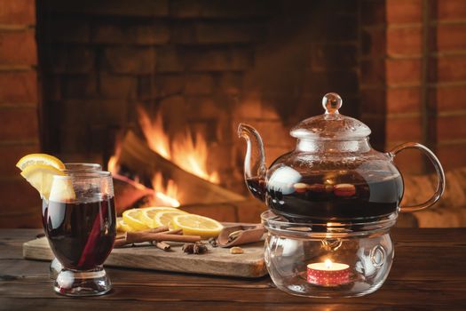 Two glasses of mulled wine and teapot on a wooden table in front of a burning fireplace.