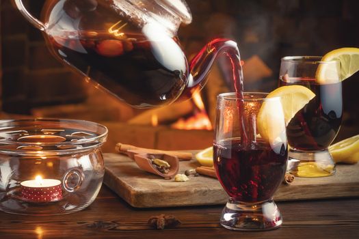 Pouring mulled wine into glasses from a teapot in a room with a fireplace.