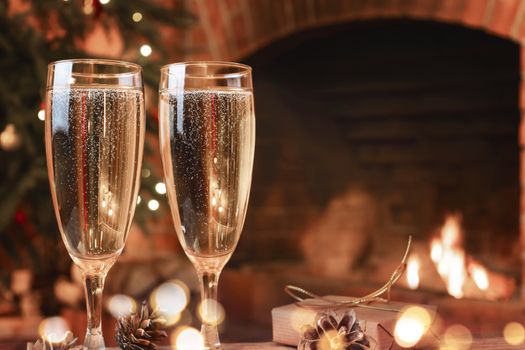 Two glasses with champagne on a wooden table in a room with a burning fireplace.