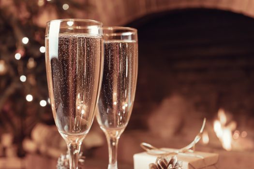 Two glasses with champagne on a wooden table in a room with a burning fireplace.