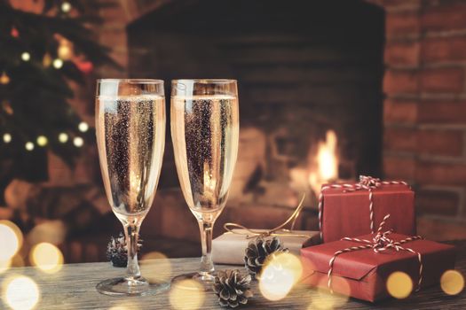 Two glasses with champagne and gifts on a wooden table in a room with a burning fireplace.