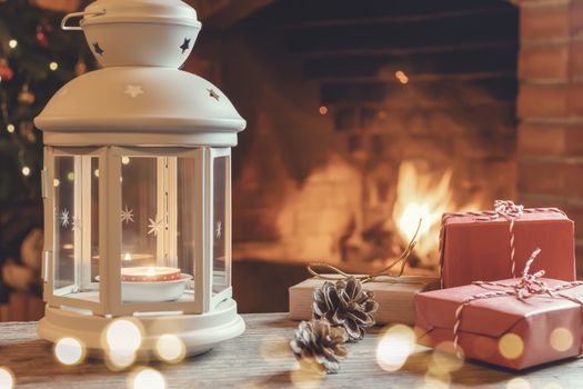 Lantern with a burning candle, gifts on a wooden table in a room with a Christmas tree and a fireplace on Christmas Eve.