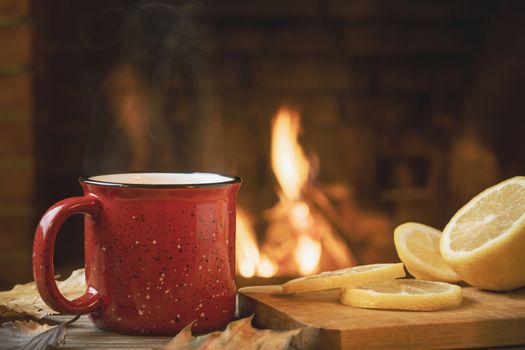 Red mug with hot tea with lemon in front of a burning fireplace, comfort and warmth of the hearth concept.