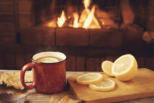 Red mug with hot tea with lemon in front of a burning fireplace, comfort and warmth of the hearth concept.