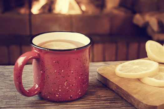 Red mug with hot tea with lemon in front of a burning fireplace, comfort and warmth of the hearth concept.