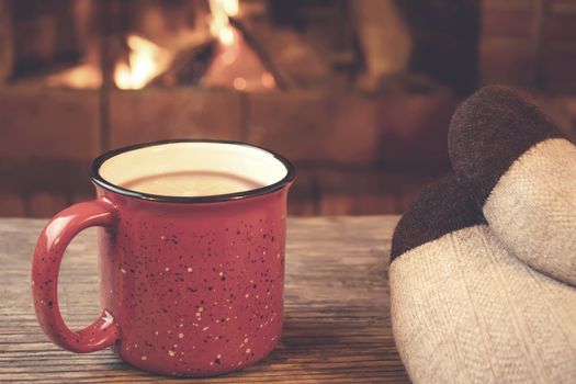 Feet in woolen socks, a red mug with hot tea opposite the burning fireplace, comfort, and warmth of the hearth concept.