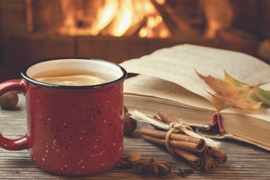 Red mug with hot tea and an open book in front of a burning fireplace, comfort, relaxation and warmth of the hearth concept.
