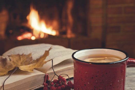 Red mug with hot tea and an open book in front of a burning fireplace, comfort, relaxation and warmth of the hearth concept.